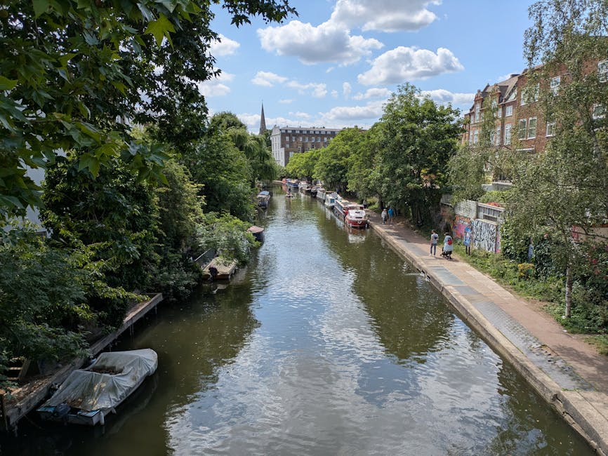A daytime view of a narrow canal with calm water reflecting the partly cloudy sky above. On the left side, dense green trees extend over the water, providing shade to a small boat docked along the can