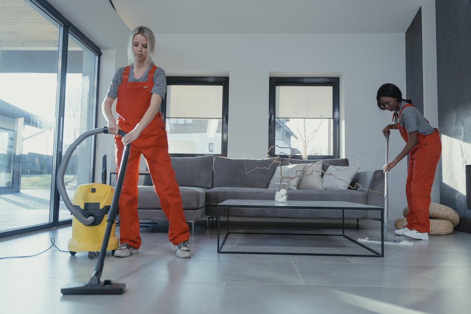 Two professional cleaners from Hackney Cleaner, dressed in grey shirts and orange overalls, are performing surface cleaning in a modern living room. The cleaner on the left is operating a yellow vacuum cleaner to clean the tiled floor, while the cleaner on the right is using a mop to clean the area around a grey sofa with light-colored cushions. The room features large windows allowing natural light, with a glass sliding door on the left. The tiled floor appears clean and shiny, and the overall space is tidy, reflecting thorough domestic cleaning suitable for end of tenancy purposes. The atmosphere emphasizes hygiene and maintenance of the living space.