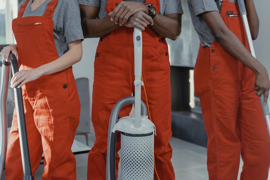 Close-up view of three professional cleaners wearing orange overalls and grey t-shirts, standing side by side in a modern interior space. The central cleaner holds a portable vacuum cleaner with a white and grey perforated container, connected by a flexible hose and a telescopic handle. The other two cleaners, positioned on either side, hold additional cleaning tools, such as hoses and spray bottles. The room features a tiled floor, smooth surfaces, and a neutral-colored wall, indicating a clean and well-maintained environment suitable for surface cleaning or deep cleaning services. Bright lighting highlights the neatness and readiness for sanitisation procedures, with all surfaces appearing spotless and dust-free, demonstrating thorough hygiene and cleaning preparation. The image is associated with a professional cleaning service provided by Hackney Cleaner, specializing in end of tenancy cleaning, domestic cleaning, and sanitisation.