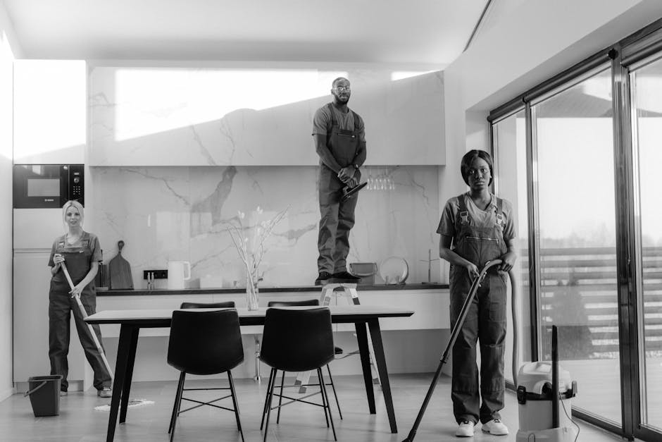 Two professional cleaners from Hackney Cleaner, dressed in grey shirts and orange overalls, are performing surface cleaning in a modern living room. The cleaner on the left is operating a yellow vacuum cleaner to clean the tiled floor, while the cleaner on the right is using a mop to clean the area around a grey sofa with light-colored cushions. The room features large windows allowing natural light, with a glass sliding door on the left. The tiled floor appears clean and shiny, and the overall space is tidy, reflecting thorough domestic cleaning suitable for end of tenancy purposes. The atmosphere emphasizes hygiene and maintenance of the living space.