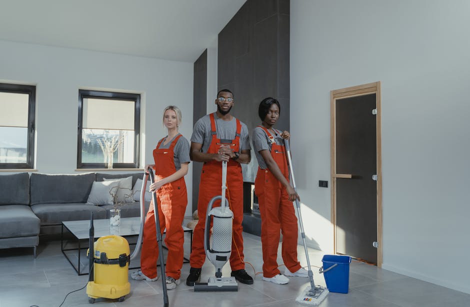 A young woman in a grey t-shirt and red work overalls holding a vacuum cleaner hose, standing in a well-lit modern room with large windows and white walls. Behind her, a male cleaner in similar attire
