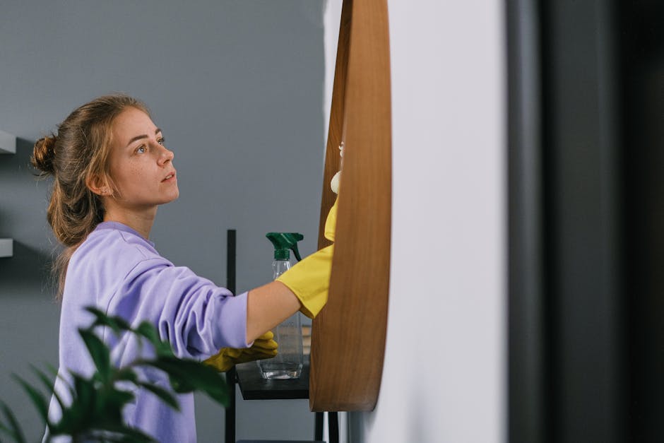 A young woman with light brown hair tied in a bun is engaged in surface cleaning in a modern interior. She is wearing a lavender-colored sweatshirt and yellow rubber gloves, and is holding a green spray bottle in her right hand while wiping a wooden surface with a cloth. The background features a grey wall and a small shelf with cleaning supplies, indicating a domestic or commercial cleaning setting. The wooden surface appears well-maintained and clean, with a shiny finish reflecting controlled lighting. The overall scene conveys thorough sanitisation and deep cleaning, consistent with the offerings of Hackney Cleaner for Hoxton flat cleaning tips near Hackney Empire, HACKNEY Cleaner.