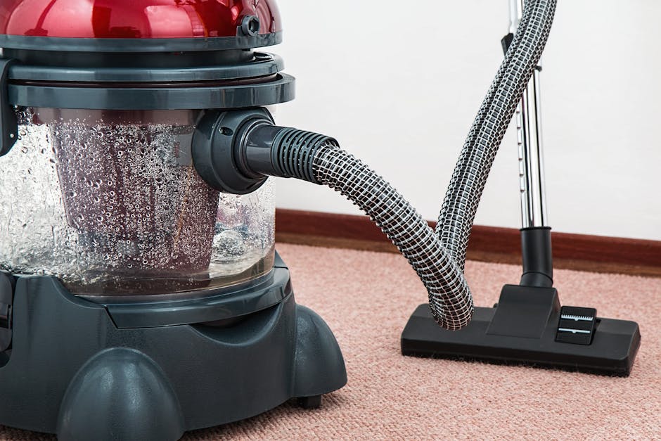 Close-up of a vacuum cleaner with a transparent dust container filled with debris, connected to a flexible, black-and-white patterned hose attached to a floor cleaning head, situated on a pinkish carpet in a residential room. The vacuum cleaner is positioned near a white wall with a wooden skirting board, and the scene is well-lit, emphasizing the cleanliness and maintenance of the carpet surface. The image illustrates surface cleaning and deep cleaning processes, consistent with professional domestic cleaning services provided by Hackney Cleaner, as featured on the Mare Street E8 carpet cleaning specialists page.