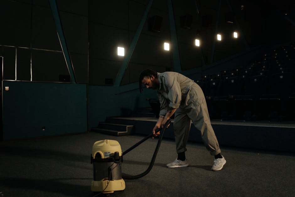 A person using a vacuum cleaner on a dark carpeted floor in an auditorium or theatre, with blue walls and tiered seating in the background. The individual is dressed in casual grey clothing and white sneakers, focusing on surface cleaning. The lighting is dim, with some overhead lights illuminating the area, demonstrating a deep cleaning or maintenance process as part of domestic or commercial cleaning services, with Hackney Cleaner specializing in professional hygiene and surface sanitation.