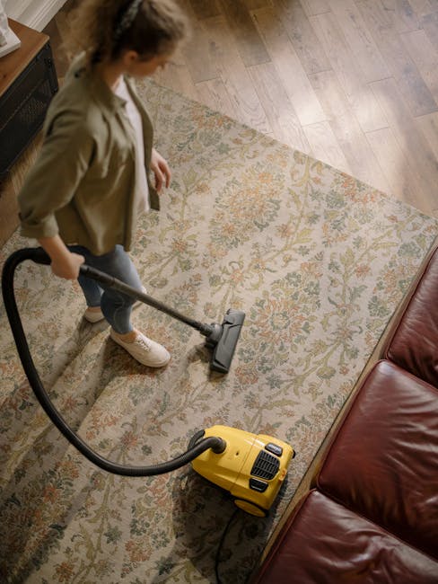Close-up of a vacuum cleaner with a transparent dust container filled with debris, connected to a flexible, black-and-white patterned hose attached to a floor cleaning head, situated on a pinkish carpet in a residential room. The vacuum cleaner is positioned near a white wall with a wooden skirting board, and the scene is well-lit, emphasizing the cleanliness and maintenance of the carpet surface. The image illustrates surface cleaning and deep cleaning processes, consistent with professional domestic cleaning services provided by Hackney Cleaner, as featured on the Mare Street E8 carpet cleaning specialists page.
